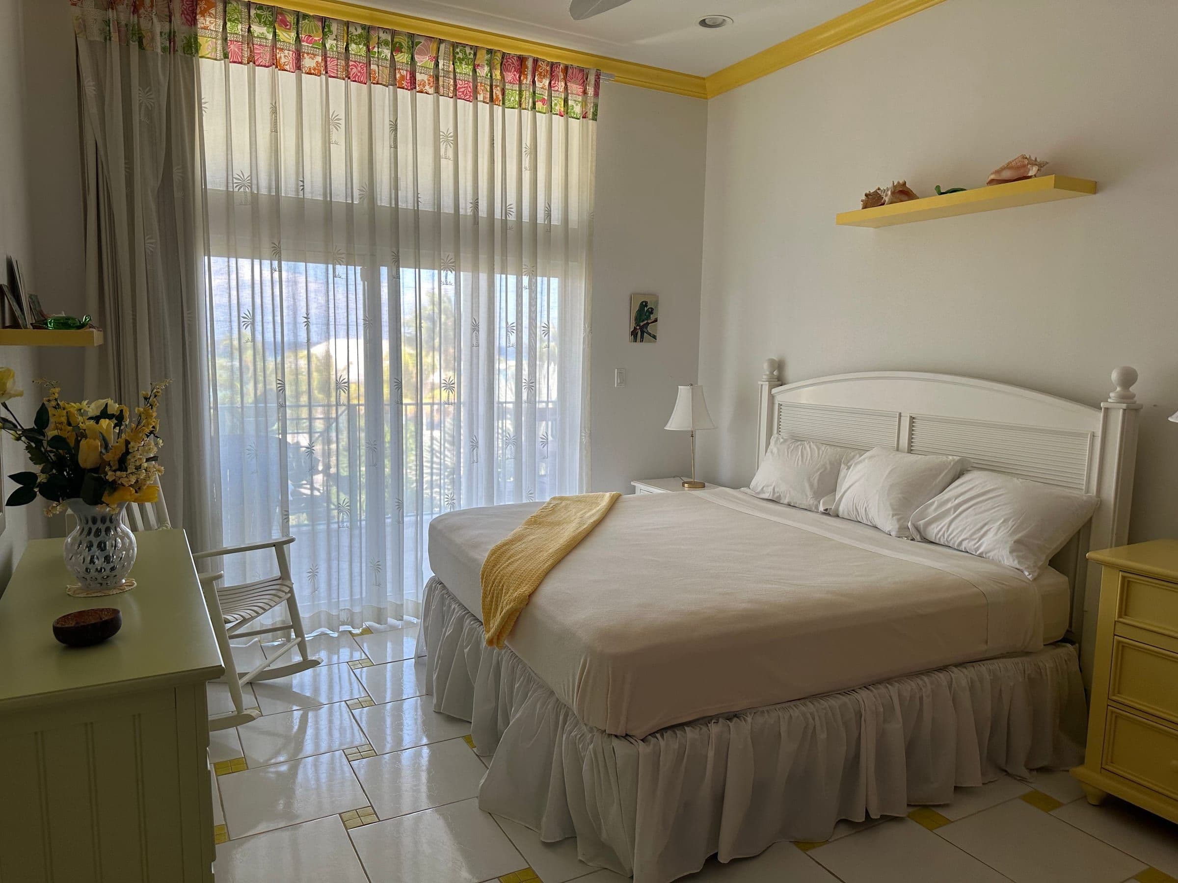 The third bedroom. Queen bed, yellow trim, sheer curtains, rocking chair.